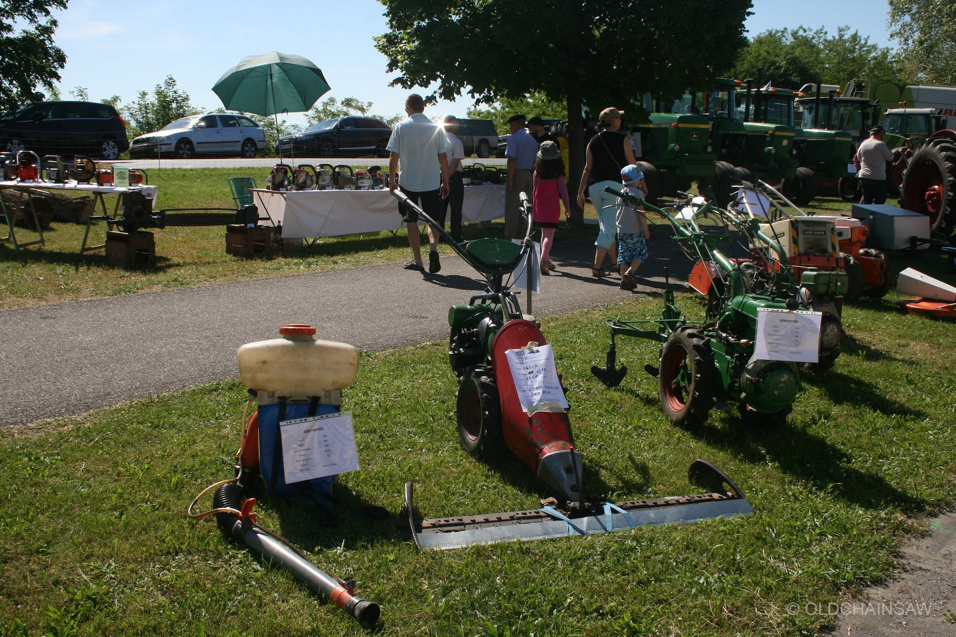 Matériel agricole ancien exposé au premier plan, avec le stand de tronçonneuses OLDCHAINSAW visible à l’arrière-plan parmi les différentes présentations de la journée.
